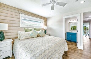 Un dormitorio con cama, mesita de noche, ventilador de techo, ventana con persianas y un baño visible a través de una puerta abierta.