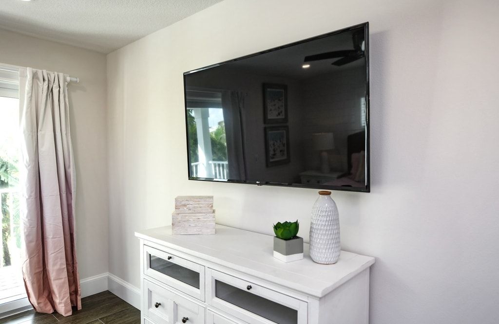 A room with a flat-screen TV mounted on the wall, a white dresser with drawers, a small plant, and a decorative vase.