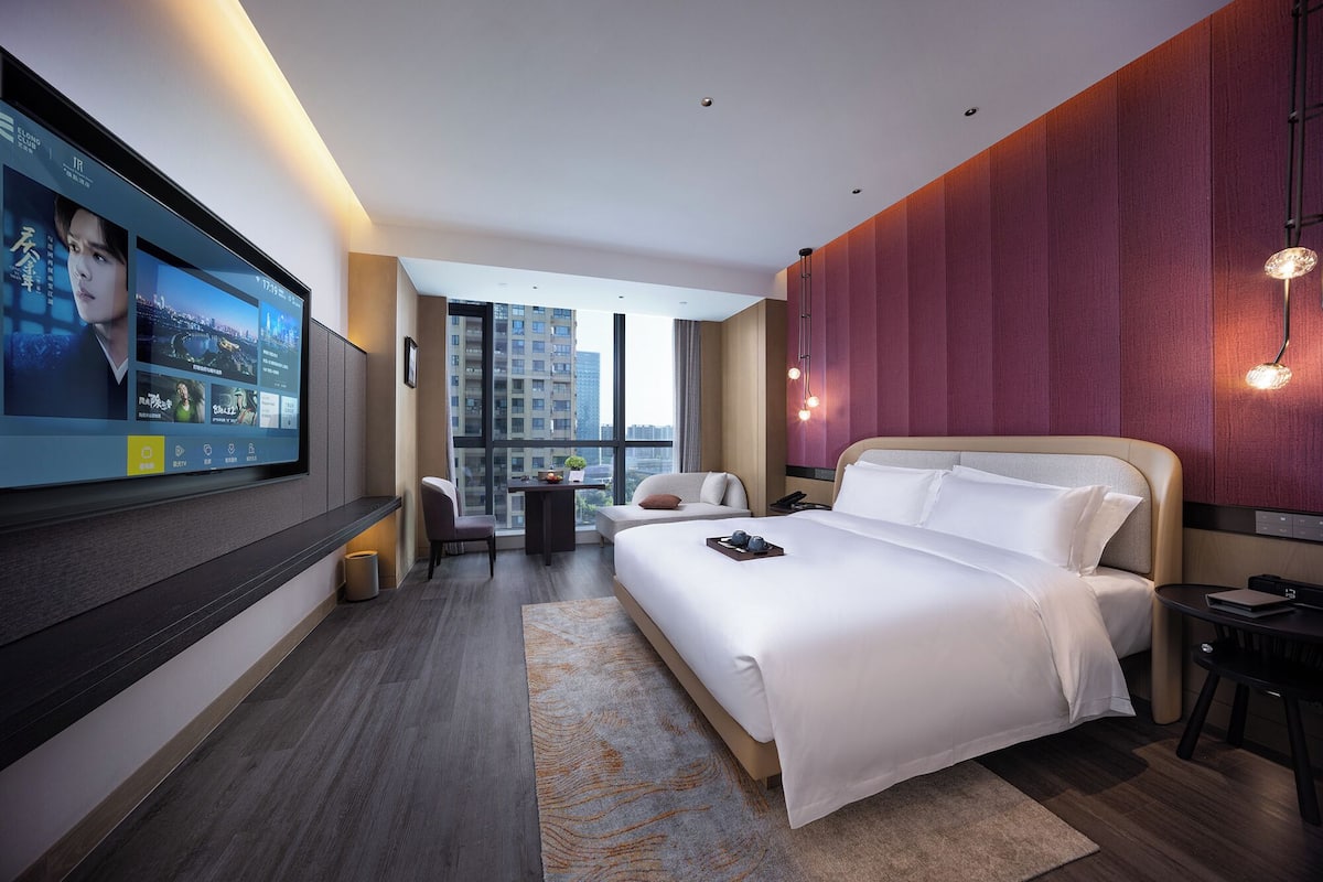 Ein modernes Hotelzimmer mit großem Fernseher, einem Bett, einem Schreibtisch und Blick auf die Stadt.