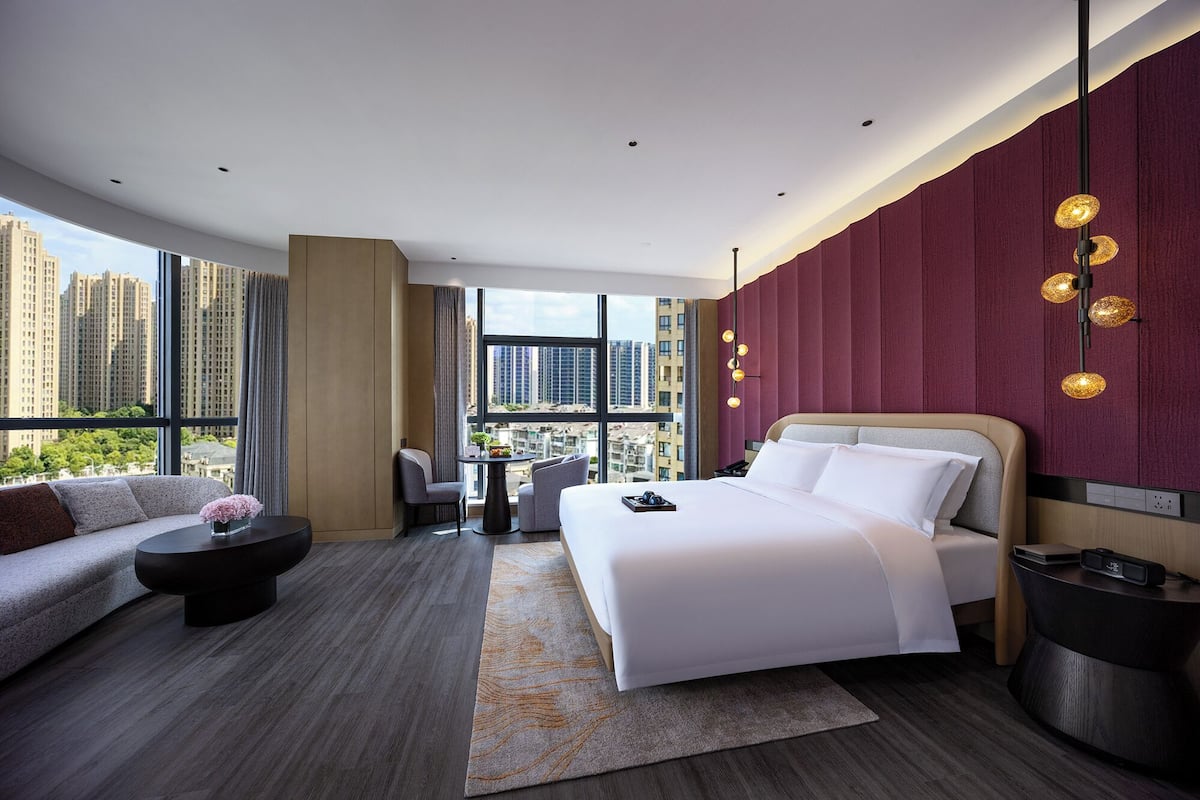 Ein modernes Hotelzimmer mit einem großen Bett, einer Couch, einem Esstisch und Blick auf hohe Gebäude.