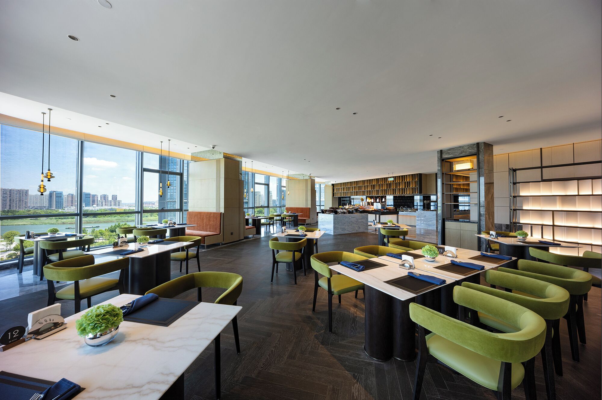 A modern restaurant with high ceilings and large windows offering city views. Tables are arranged with green chairs, and a bar area is visible in the background.