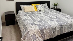 Ein Schlafzimmer mit Bett, Nachttisch, Ablagefläche, Wandregal, gerahmtem Zitat und Fenster mit Jalousien.