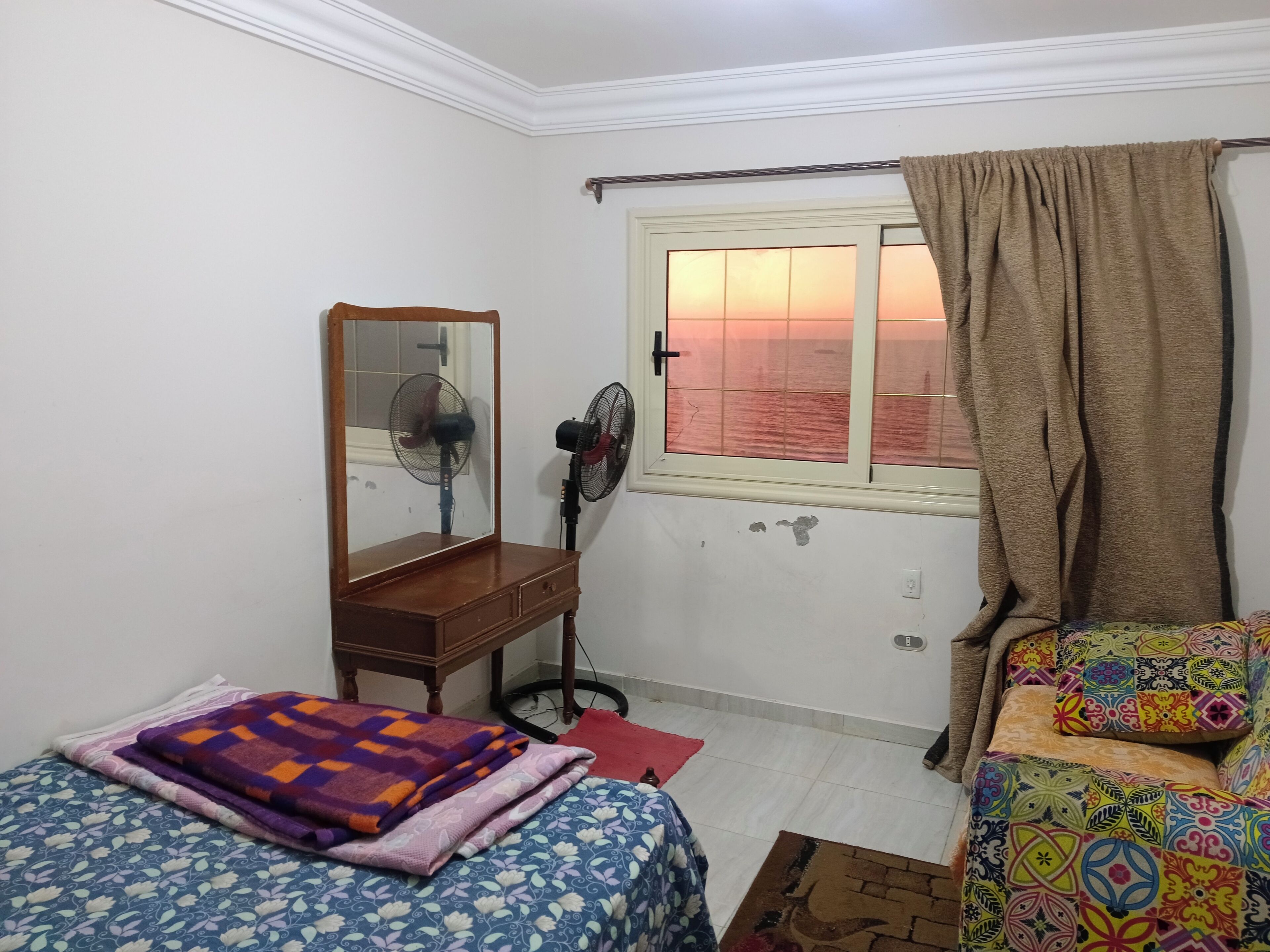 Une chambre avec deux lits, une commode en bois, un miroir, une fenêtre avec des rideaux et un ventilateur fixé au mur.