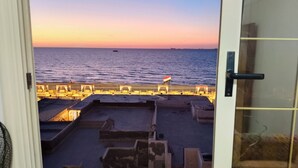 Vista desde una ventana sobre una playa con una bandera, un muelle y un barco en la distancia.