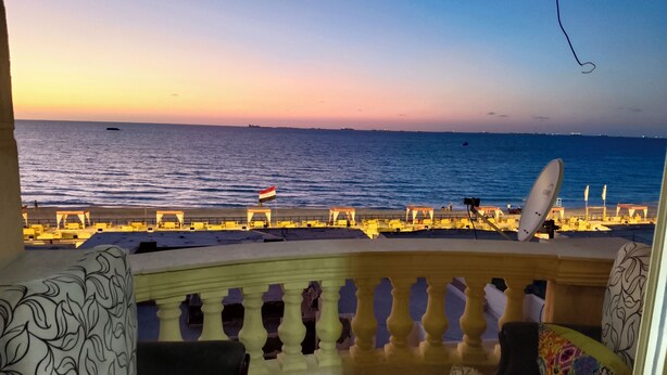 Un balcón con vistas al mar al atardecer, con un sofá, cojines de colores y una antena parabólica.