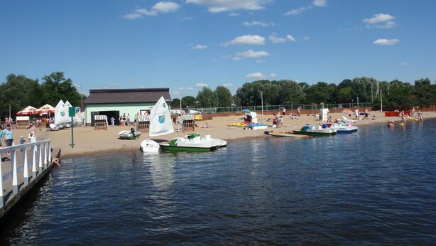 A lakeside beach with boats, a pier, and a building with a green roof. - Holiday Flat in Stepnica for 7 Persons (Stepnica)