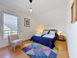 Un dormitorio con cama, una silla, una alfombra colorida, una mesita de noche con lámpara y un cuadro en la pared.