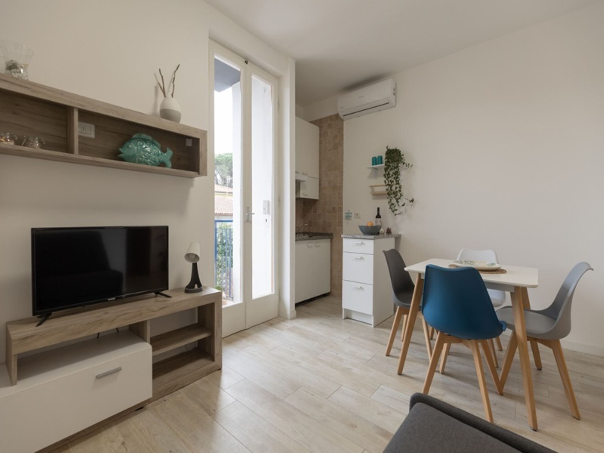 Un salon moderne doté d’un téléviseur à écran plat, d’un meuble de rangement en bois et d’un coin repas avec une table blanche et des chaises bleues.