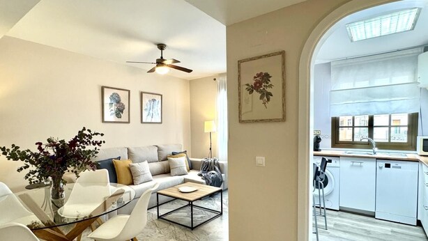 Ein modernes Wohnzimmer mit einem Glastisch, weißen Stühlen, einer Couch und einem Deckenventilator. Durch einen Bogen ist eine Küche erkennbar, ausgestattet mit weißen Schränken und einem Fenster.