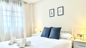 Ein ordentlich bezogenes Bett mit weißen Leinen und zwei Kissen, ein Nachttisch mit Lampe sowie ein Fenster mit transparenten Vorhängen.