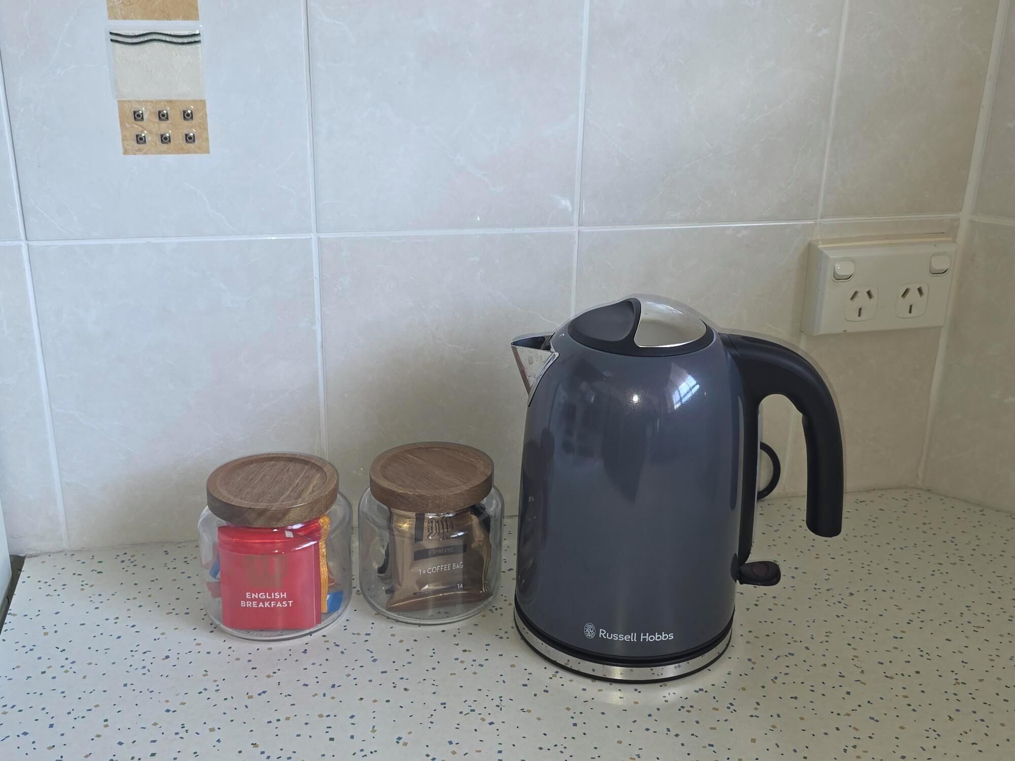 Una encimera de cocina con un hervidor, un tarro etiquetado como 'English Breakfast' y un recipiente transparente con una bolsa de café.