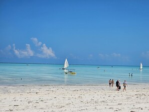 Ein Strand mit klarem türkisblauem Wasser, einigen Segelbooten und Menschen, die am Sand entlanggehen.
