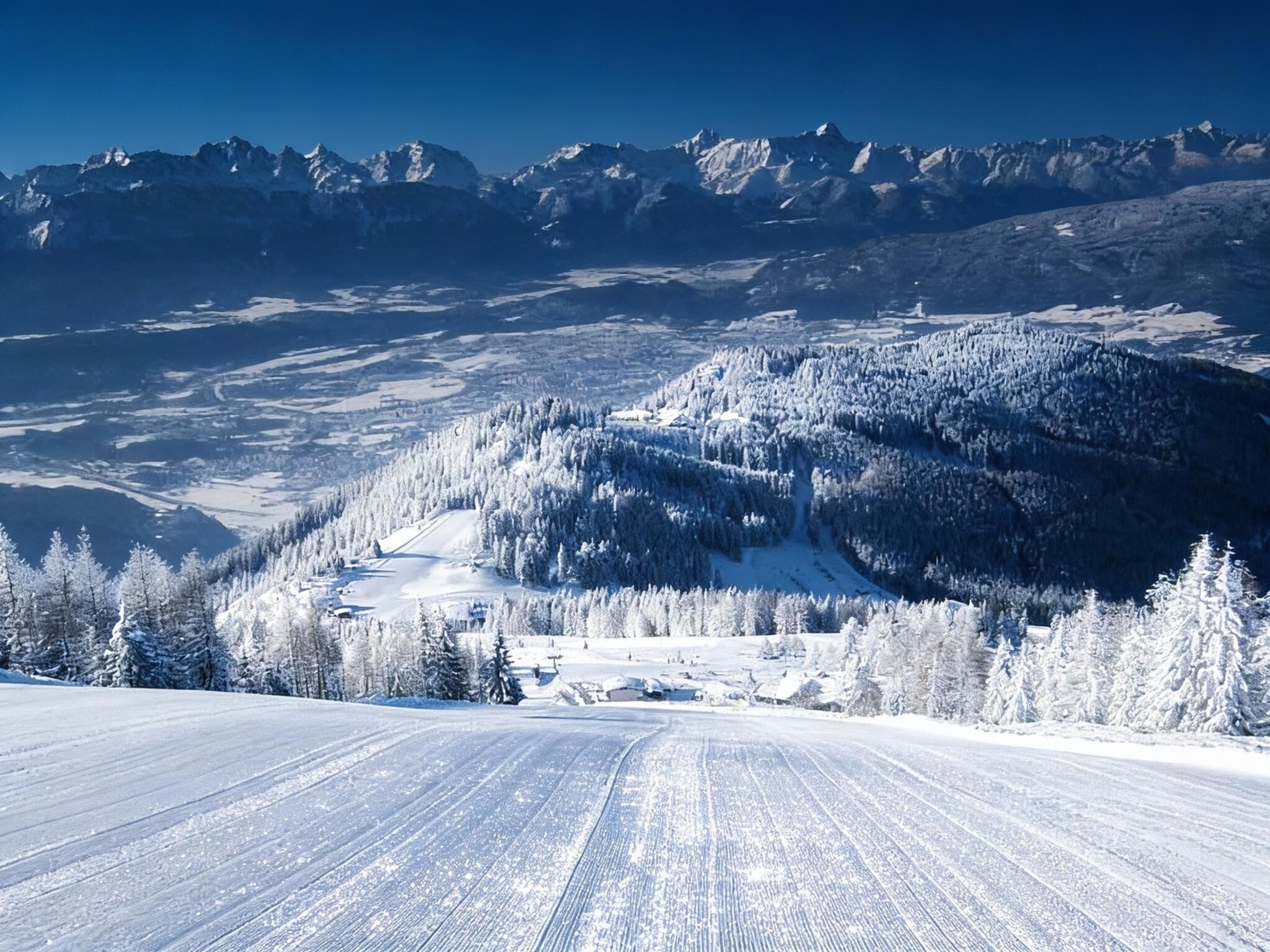 A panoramic view of a snow-covered mountain landscape with a ski slope in the foreground and a range of mountains in the background.