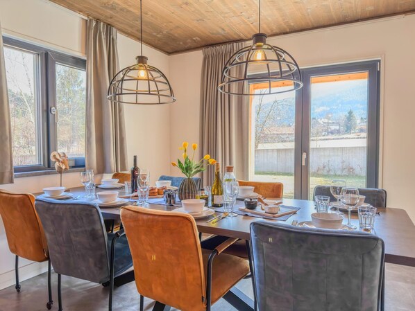 Un espace repas avec une longue table dressée pour le repas, deux lampes suspendues et une vue sur l’extérieur.