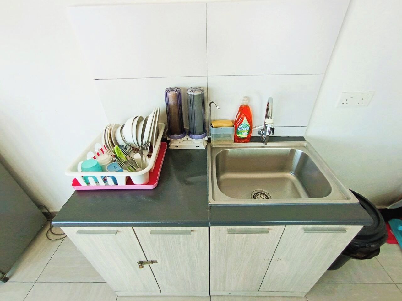 Une cuisine équipée d’un évier, d’un égouttoir, d’une bouteille de liquide vaisselle et d’une poubelle.