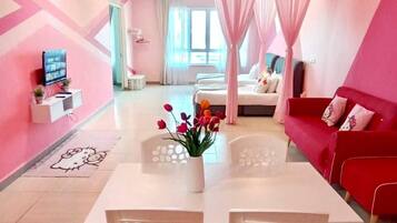 A modern hotel room with a pink and white color scheme, a dining area with a white table and chairs, a red sofa, and a large window with sheer curtains.