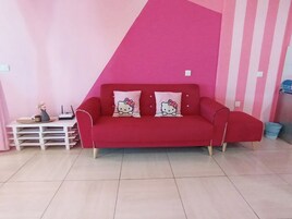 Un sofá rojo con almohadas de Hello Kitty, una mesita de noche blanca y una pared rosa con un estampado diagonal a rayas.