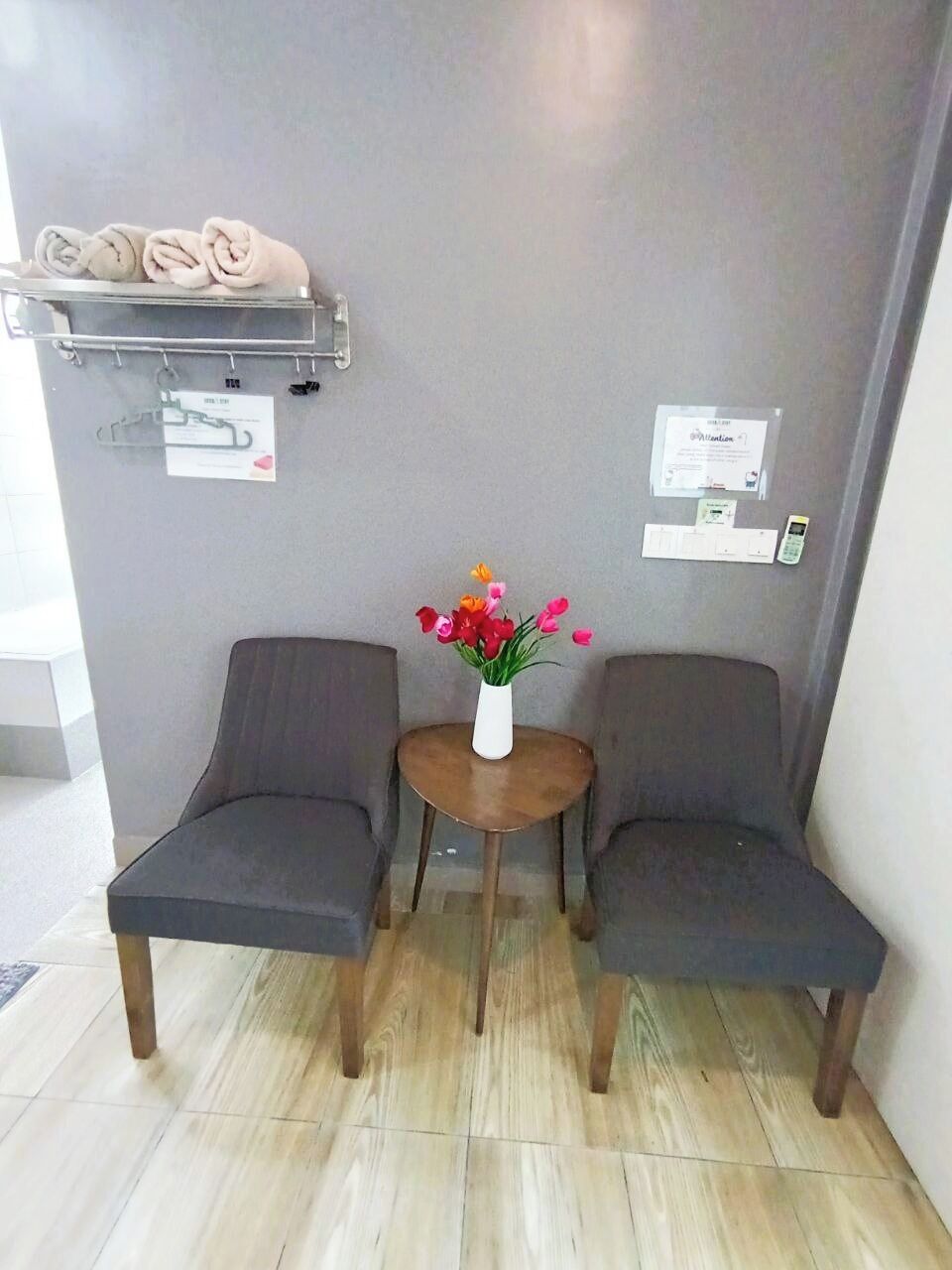 A room with two dark-colored chairs, a small round table with a vase of flowers, a towel rack with folded towels, and a wall-mounted phone.