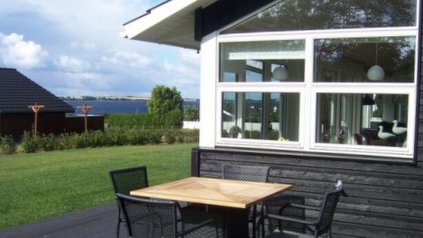 Une maison moderne dotée d’une grande fenêtre, d’une terrasse avec une table et des chaises, et offrant une vue sur un plan d’eau.