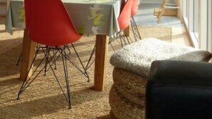 Un coin repas avec une table dressée pour deux personnes, un enfant qui joue avec un jouet, et une cuisine en arrière-plan.