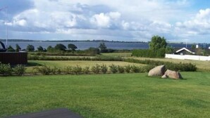 Un espace extérieur verdoyant, avec un grand rocher, une terrasse en bois et une vue sur une étendue d’eau et des bâtiments éloignés.