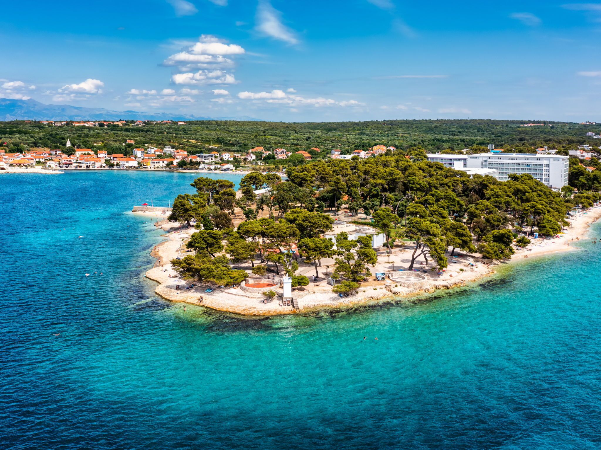 Vue aérienne d’une zone côtière avec une petite île couverte d’une végétation luxuriante, une plage de sable et une mer d’un bleu limpide.