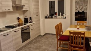 Una cocina compacta con muebles blancos, una mesa de comedor con sillas y una ventana con cortinas de encaje.