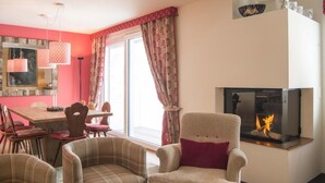 Ein Wohnzimmer mit Kamin, Karo-Sesseln, einem Essbereich mit rotem Tisch und einem großen Fenster mit Vorhängen.