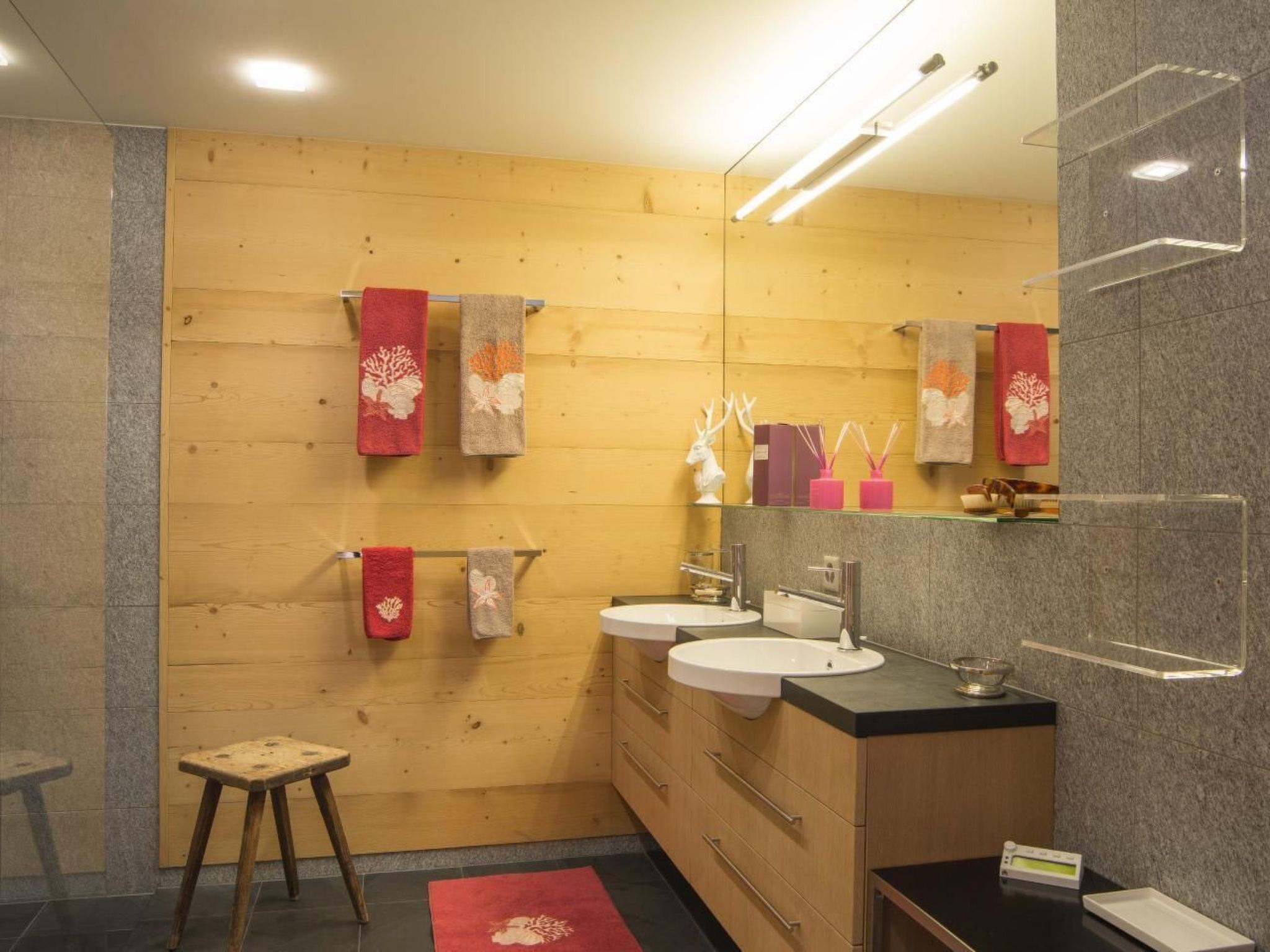 A modern bathroom with wooden walls, two sinks, a mirror, and a stool.