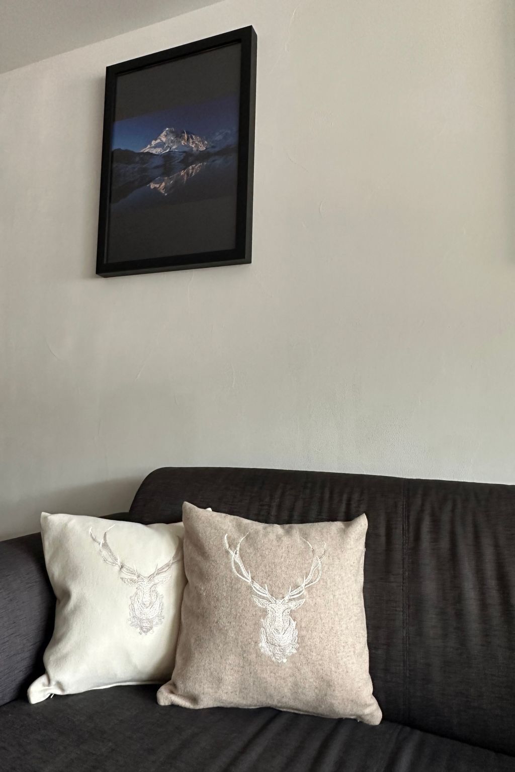 Ein dunkelgraues Sofa mit zwei dekorativen Kissen mit einem Hirschmuster. Darüber an der Wand hängt ein gerahmtes Bild einer Berglandschaft.
