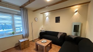 Una sala de estar acogedora con suelo de madera, un sofá negro, una pequeña mesa de madera y una ventana con cortinas.
