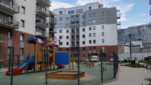 Un terrain de jeux délimité par une clôture, équipé d’un toboggan et de structures d’escalade, entouré d’un bâtiment en briques et d’un immeuble d’appartements moderne.