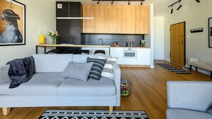 Un salon moderne avec un canapé gris, une table à manger en bois et une cuisine dotée de meubles en bois et d’un réfrigérateur noir.