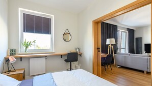 Une chambre moderne avec un grand lit, un bureau avec une chaise, une petite table, une plante en pot et une fenêtre avec des stores.