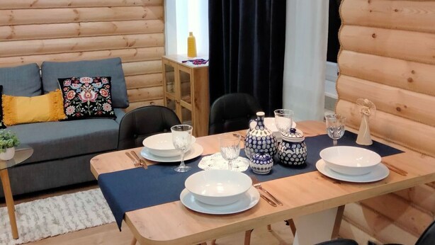Een eetgedeelte met een houten tafel voor vier personen, omringd door zwarte stoelen. Een houten bank met een bloemkussen staat tegen de muur, en erboven hangt een flatscreen-televisie.