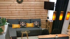 Een gezellige woonkamer met een houten wand, een grijze bank met gele kussens, een klein rond tafeltje, en een eethoek met een houten tafel en zwarte stoelen.