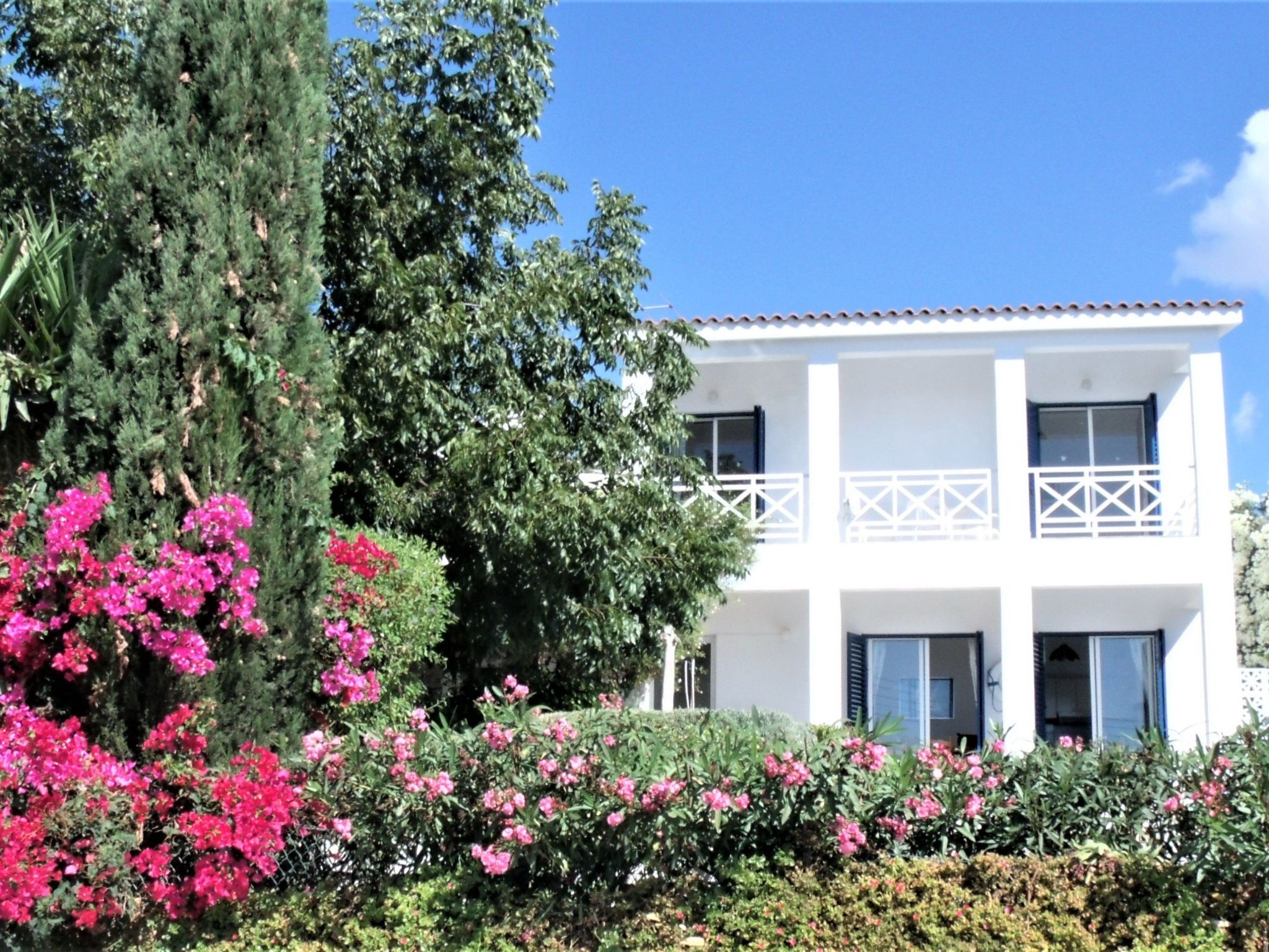 A two-story white building with balconies, surrounded by lush greenery and pink flowers.
