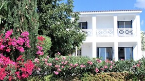 Un edificio blanco de dos pisos con balcones, rodeado de exuberante follaje y flores rosadas.