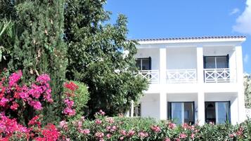 A two-story white building with balconies, surrounded by lush greenery and pink flowers.