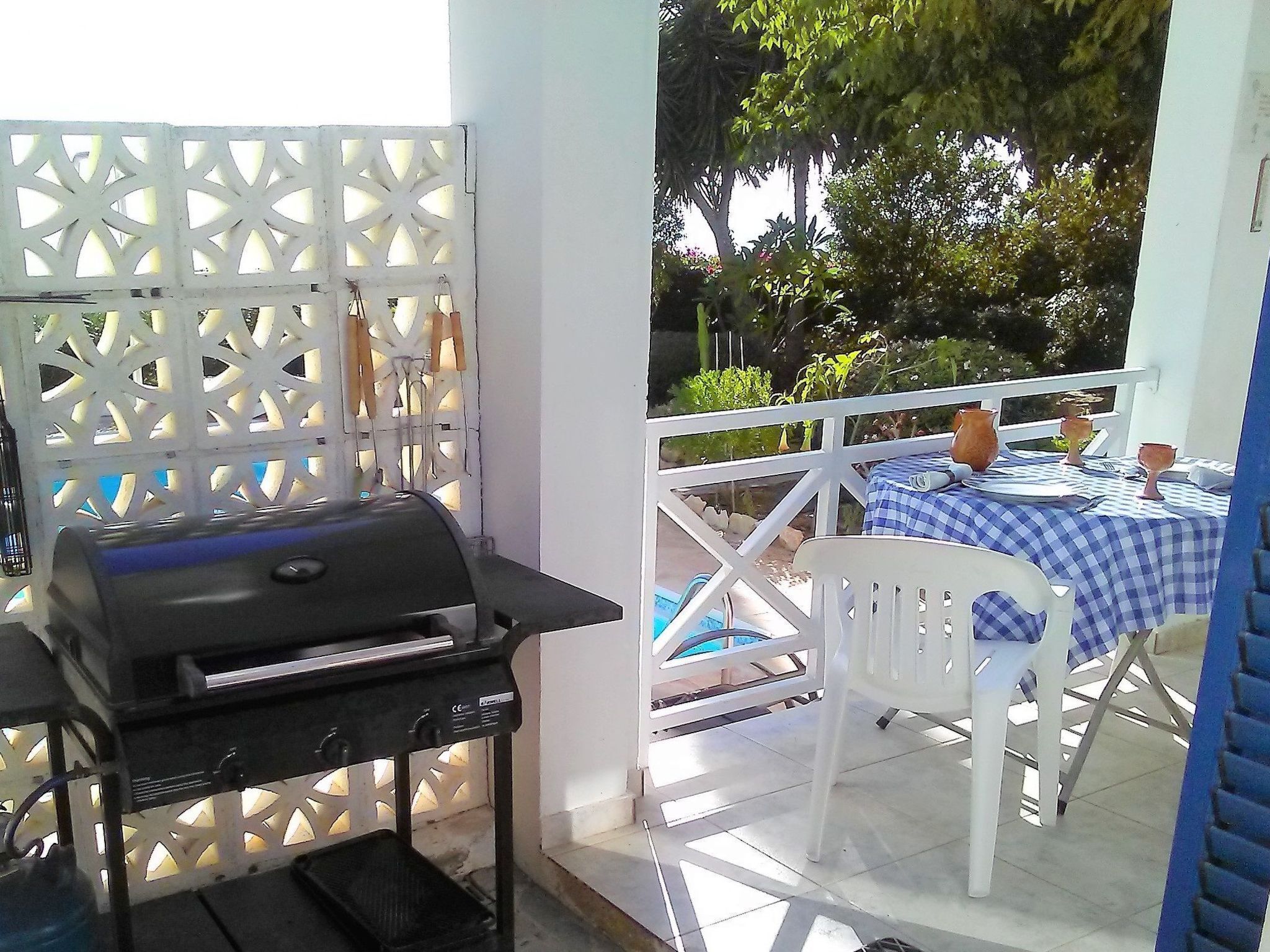 Een terras buiten met een barbecue, witte stoelen en een geruit tafelkleed.