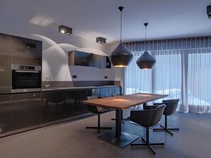 Une cuisine moderne dotée d’un four encastré, d’un coin repas avec une table en bois et des chaises noires, et d’un éclairage suspendu.