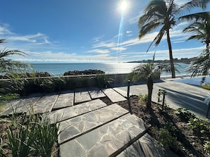 Ein steiniger Weg führt zu einem Poolbereich mit Palmen und Blick auf das Meer.
