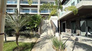Un bâtiment moderne doté de nombreux balcons, d’un escalier central et entouré de verdure, notamment de palmiers et d’autres plantes.