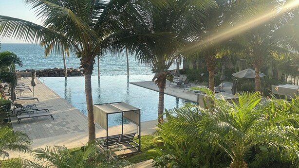 Un espace aménagé au bord de la piscine, avec des chaises longues, un coin salon abrité et des palmiers.