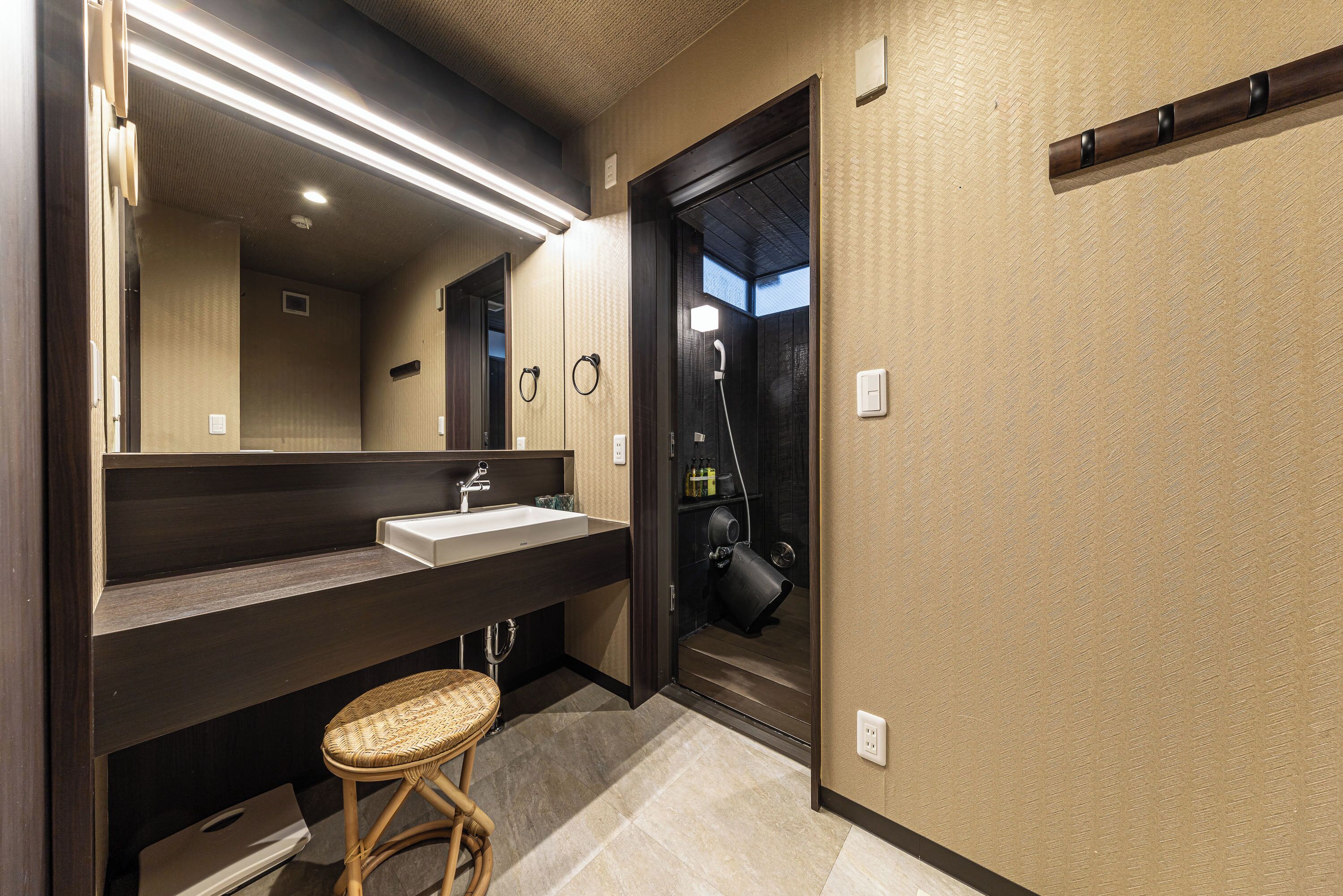 Une salle de bain moderne avec un grand miroir, un meuble-lavabo en bois foncé et un tabouret en osier.