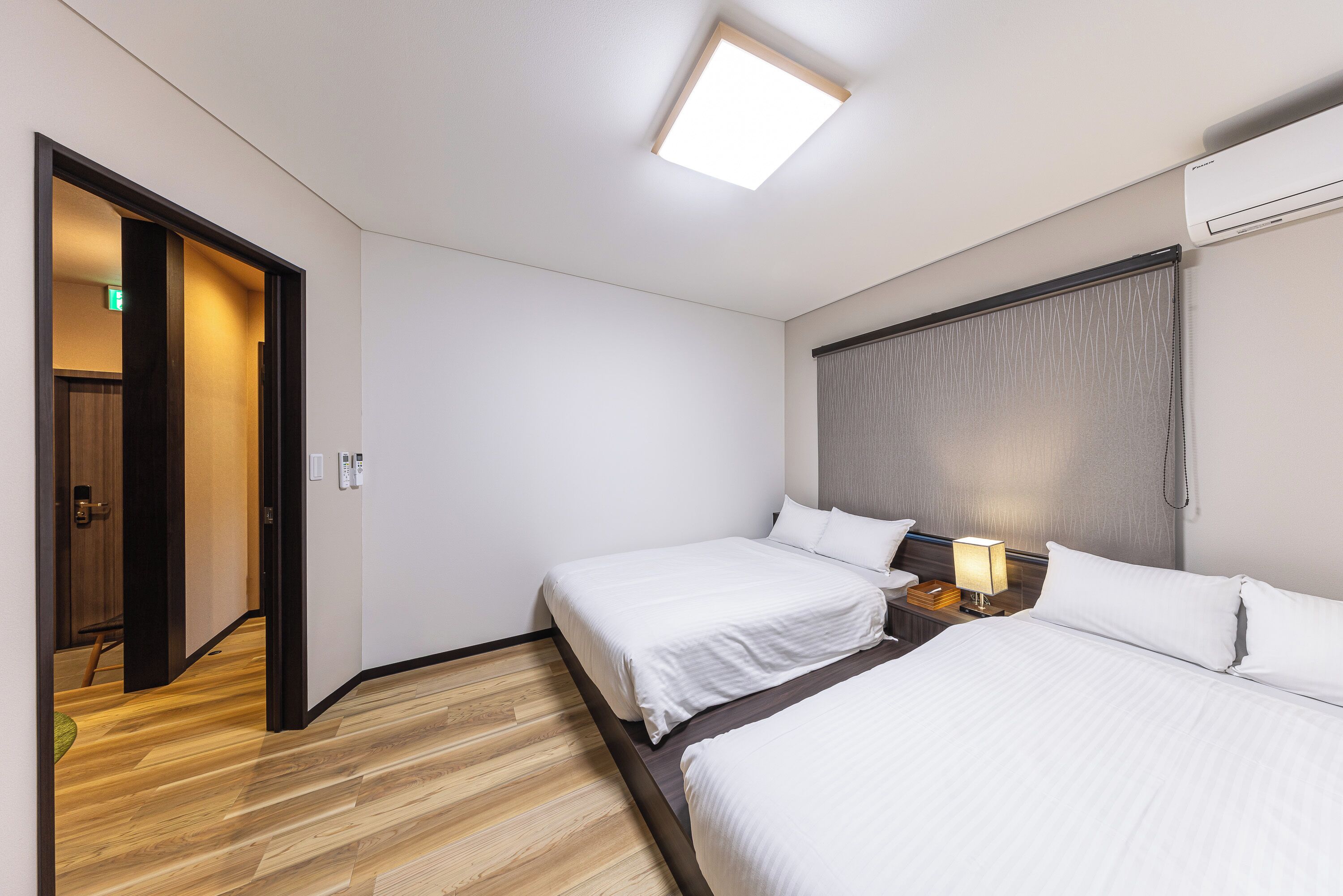 Habitación de hotel con dos camas, suelo de madera, luz de techo, equipo de aire acondicionado y mesita de noche con lámpara.