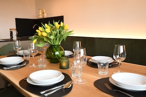 Ein Esstisch ist gedeckt mit Tellern, Besteck und Gläsern, in der Mitte steht ein Blumenstrauß mit gelben Blumen.