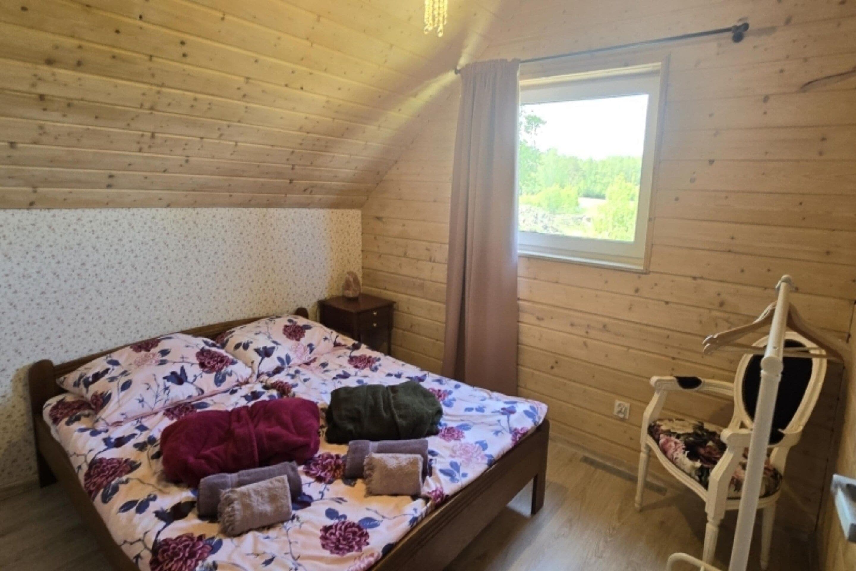 A bedroom with a wooden ceiling, a bed with floral bedding, a nightstand, a chair, and a window with a view of greenery.