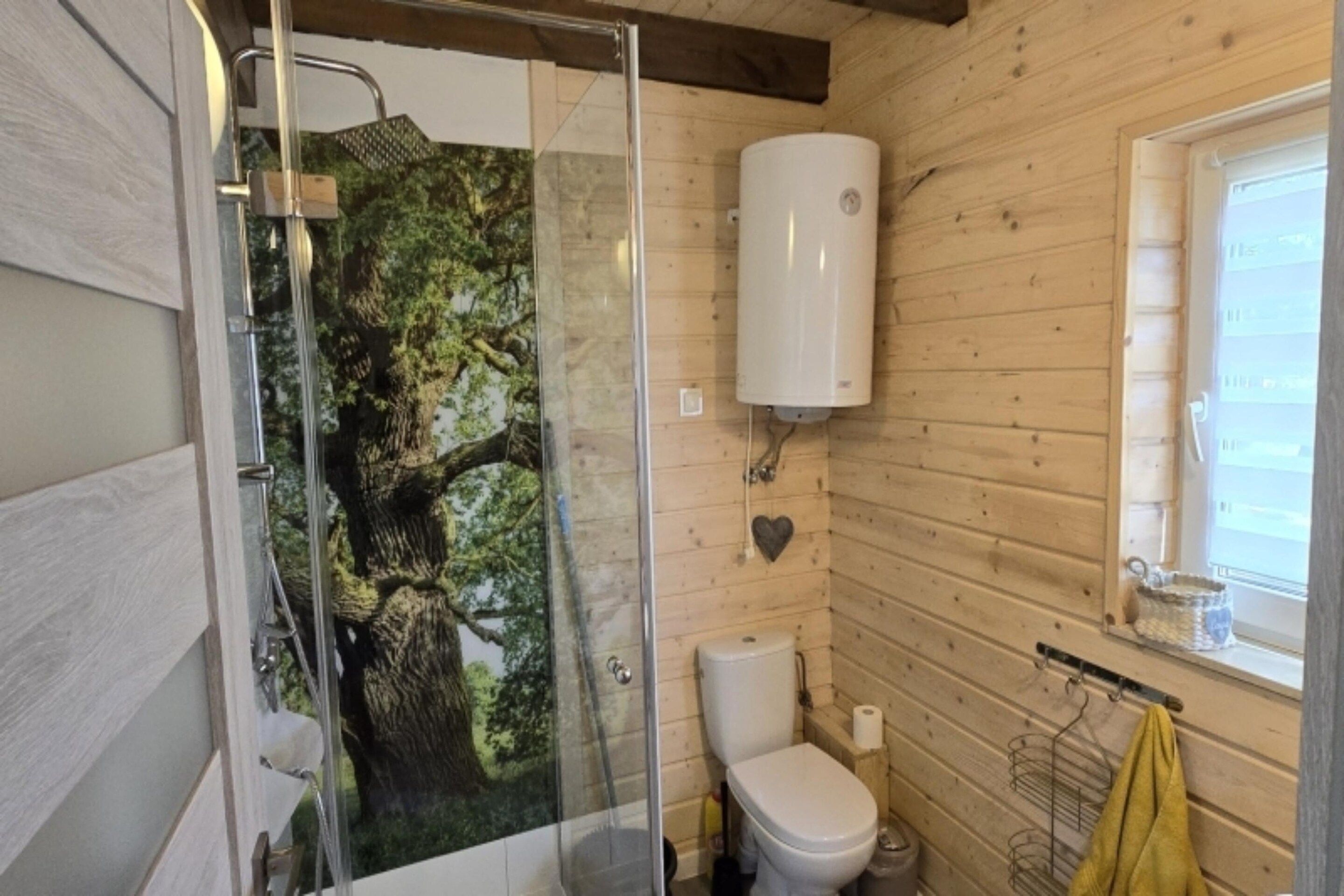 A bathroom with a glass shower enclosure, a toilet, and a window with a towel hanging on the sill.