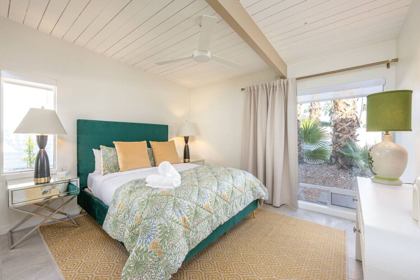 Un dormitorio con cabecera verde, edredón estampado y vistas a un jardín a través de una ventana.